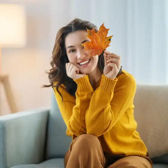 Eine Frau im gelben Pullover, die sich ein gelbes Herbstblatt vor das Auge hält