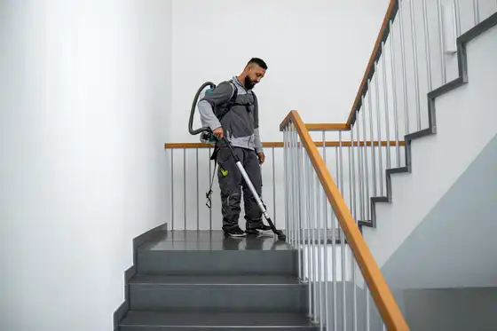 Ein Mitarbeiter des raumgut Facility Service steht in einem sehr sauberen, modernen Treppenhaus in grauer Arbeitskleidung und saugt professionell die Treppenabsätze. Das Foto vermittelt Professionalität im Bereich der Gebäudereinigung.